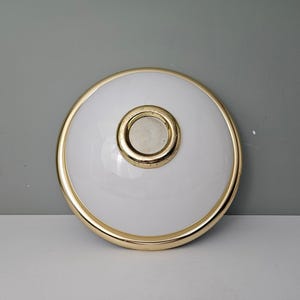 Könnte beinhalten: Eine weiße Deckenleuchte mit goldener Einfassung und einem runden, goldfarbenen Mittelteil.