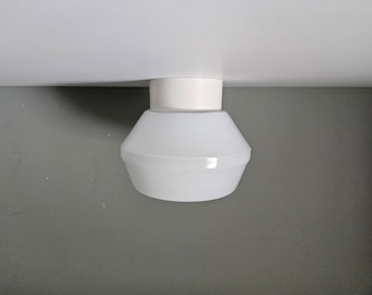 May include: A white, geometric-shaped ceiling light fixture. The light has a cylindrical base and a flared, opaque shade. The fixture is mounted on a white ceiling against a gray wall.