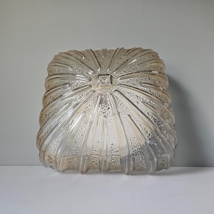 May include: A clear, textured glass light fitting with a square shape and a radiating, ridged design. The fitting has a central square detail and a slightly amber hue, suggesting vintage or antique styling.