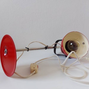 K&ouml;nnte beinhalten: Eine rote Vintage-Schreibtischlampe mit rundem Sockel und schwenkbarem Arm. Die Lampe hat einen roten Lampenschirm und einen Chromarm, mit wei&szlig;em Kabel und Schalter. Die Lampe ist f&uuml;r einen Schreibtisch oder Tisch konzipiert.
