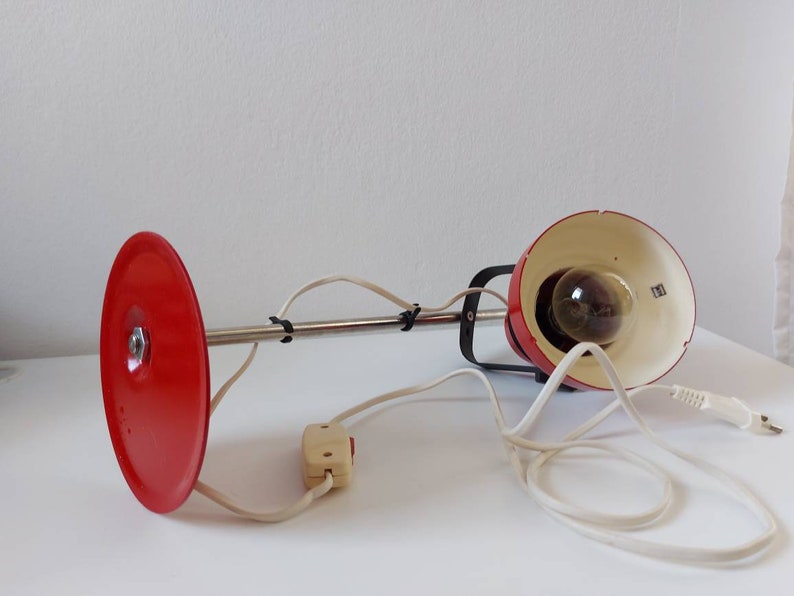 K&ouml;nnte beinhalten: Eine rote Vintage-Schreibtischlampe mit rundem Sockel und schwenkbarem Arm. Die Lampe hat einen roten Lampenschirm und einen Chromarm, mit wei&szlig;em Kabel und Schalter. Die Lampe ist f&uuml;r einen Schreibtisch oder Tisch konzipiert.
