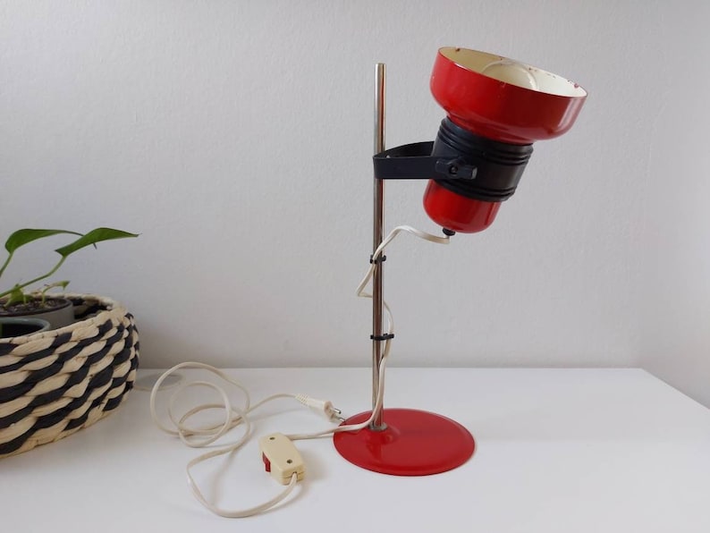 K&ouml;nnte beinhalten: Eine rote und schwarze Vintage-Schreibtischlampe mit rundem Sockel und verstellbarem Kopf. Die Lampe hat einen Chromschaft und einen wei&szlig;en Innenraum. Die Lampe ist mit einem wei&szlig;en Kabel und einem beigen Schalter an eine Steckdose angeschlossen.