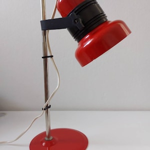 K&ouml;nnte beinhalten: Eine rot-schwarze Schreibtischlampe mit rundem Fu&szlig; und verstellbarem Kopf. Die Lampe hat einen verchromten Stiel und ein wei&szlig;es Netzkabel. Der Lampenkopf ist nach unten geneigt, und der Sockel hat einen Durchmesser von etwa 15 cm.