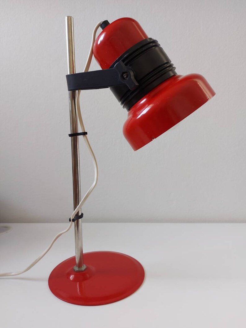 K&ouml;nnte beinhalten: Eine rot-schwarze Schreibtischlampe mit rundem Fu&szlig; und verstellbarem Kopf. Die Lampe hat einen verchromten Stiel und ein wei&szlig;es Netzkabel. Der Lampenkopf ist nach unten geneigt, und der Sockel hat einen Durchmesser von etwa 15 cm.