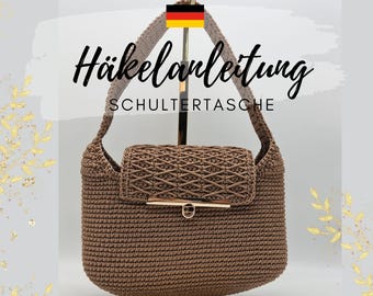 Puede incluir: Un bolso de hombro de ganchillo marrón con cierre de solapa y cierre de metal dorado. El bolso se muestra en un soporte de metal dorado. El texto "Häkelanleitung Schultertasche" es visible sobre el bolso.
