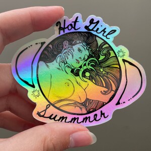 May include: Holographic sticker featuring a nude woman with long flowing hair, surrounded by a circular design. The sticker has the words "Hot Girl Summer" in black script. The sticker has a rainbow color effect.