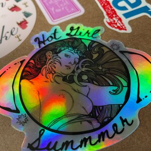 May include: Holographic sticker featuring a nude illustration of a woman with long hair, surrounded by the words "Hot Girl Summer." The sticker has a rainbow sheen and is set against a brown background, with other stickers visible.