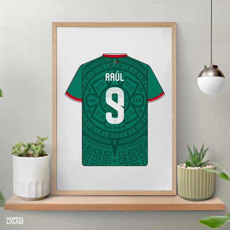 May include: A framed print featuring a green soccer jersey with a dark green Aztec-inspired circular pattern. The jersey displays the name RA&Uacute;L and the number 9 in white, with red and white trim on the sleeves and collar, presented in a light wood frame.