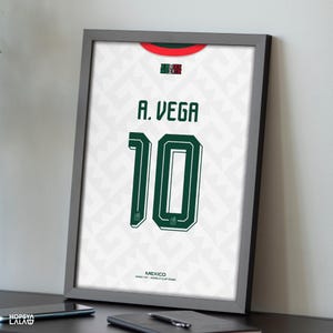 May include: A framed print of a white Mexico soccer jersey featuring a subtle geometric pattern, a red and green collar, the name A. VEGA, and the number 10 in dark green. The text MEXICO AWAY KIT WORLD CUP 2026 is printed at the bottom.