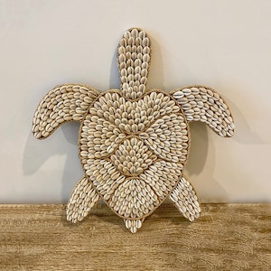 Puede incluir: Una decoración de pared con forma de tortuga hecha de materiales naturales. La tortuga está hecha de pequeñas conchas blancas y cuerda marrón.