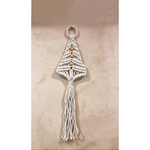 Può includere: Un'appendice da parete a forma di albero di Natale in macramè bianco con una ghirlanda di perline di legno e un lungo fiocco.
