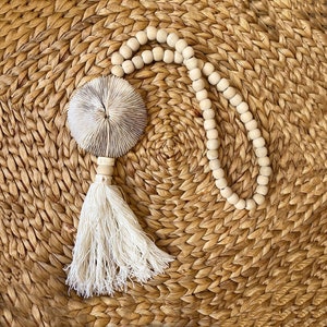 May include: A white tassel hangs from a white coral-shaped ornament. The ornament is attached to a string of white wooden beads. The beads are strung on a natural woven surface.