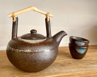 May include: A brown ceramic teapot with a bamboo handle and lid. Two small brown ceramic teacups with blue and white glaze are next to the teapot on a wooden surface.