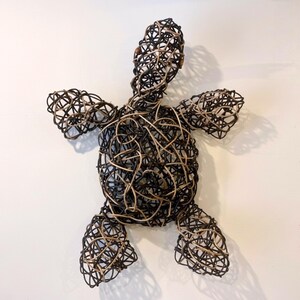 May include: A handcrafted wall art piece in the shape of a turtle. The sculpture is made of woven dark brown and tan-colored wire, creating a textured, open design. The turtle has outstretched limbs and a rounded shell.