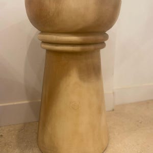 May include: A light tan wooden stool with a smooth, cylindrical base that widens toward the bottom. The top features a wide, bowl-shaped seat with a rounded rim, separated from the base by a distinct horizontal ring detail around the center.
