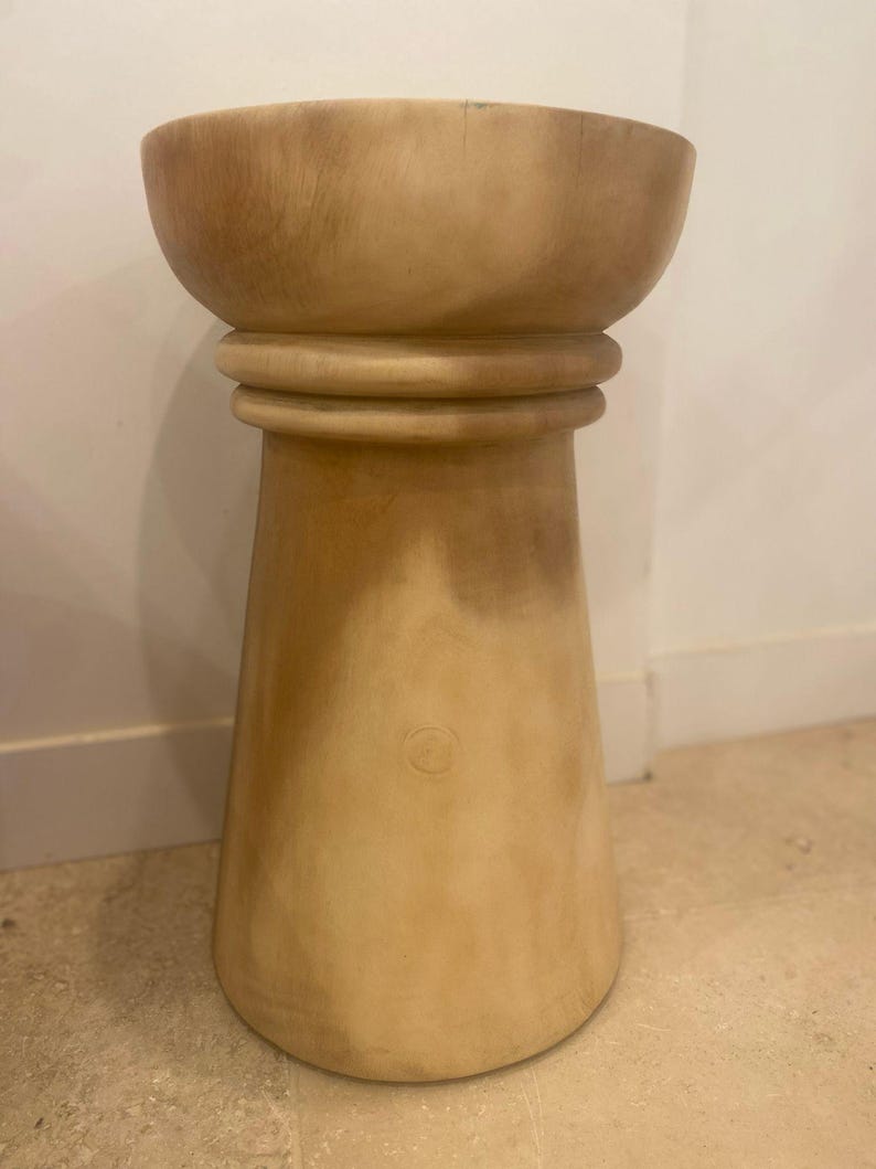 May include: A light tan wooden stool with a smooth, cylindrical base that widens toward the bottom. The top features a wide, bowl-shaped seat with a rounded rim, separated from the base by a distinct horizontal ring detail around the center.