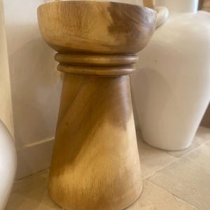 May include: A light brown wooden stool with a smooth, cylindrical top section and a wider, tapered base. The stool features a distinct horizontal ring detail separating the top bowl-like surface from the flared bottom, showcasing natural wood grain.