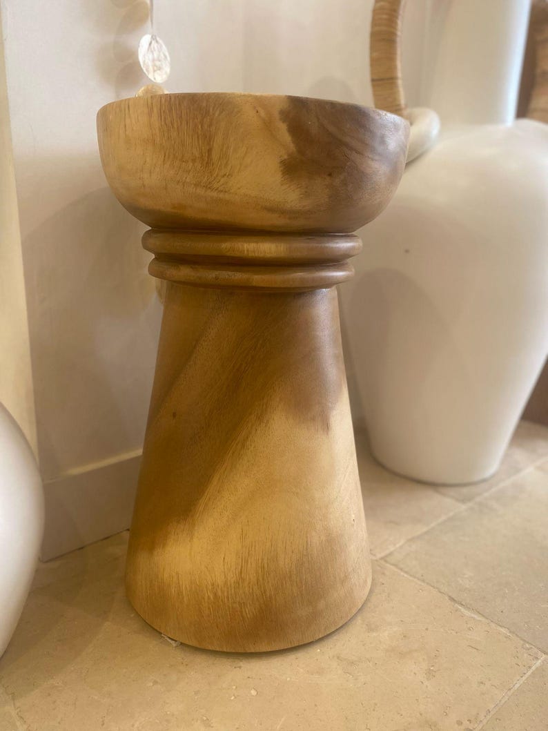 May include: A light brown wooden stool with a smooth, cylindrical top section and a wider, tapered base. The stool features a distinct horizontal ring detail separating the top bowl-like surface from the flared bottom, showcasing natural wood grain.