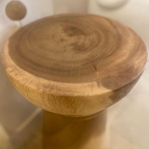 May include: A round wooden stool featuring a natural light brown finish with visible wood grain patterns on the circular top surface. The stool has a smooth, polished texture and a solid, cylindrical base that tapers slightly toward the center.