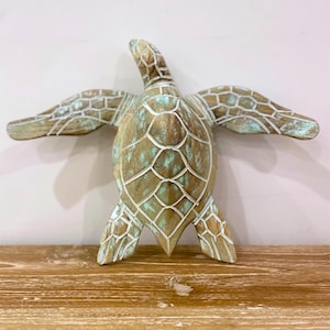 Può includere: Una scultura di tartaruga in legno con pinne spiegate. La tartaruga è dipinta in tonalità marroni e turchesi, con accenti bianchi. La scultura è esposta su una superficie di legno.