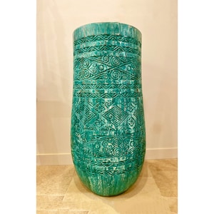 Pode incluir: Um vaso alto de cerâmica azul-esverdeado com padrões intrincados esculpidos. O vaso tem um fundo arredondado e uma parte superior ligeiramente alargada.