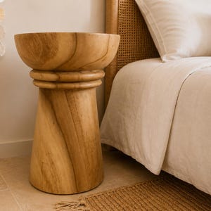 May include: A light brown wooden side table with a smooth, circular top and a tapered, cylindrical base. The table features a distinct horizontal ring detail just below the top surface, showcasing the natural wood grain patterns throughout the piece.