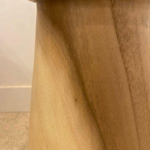 May include: A light brown wooden stool with a smooth, polished finish. The stool features a tapered, cylindrical body with a slightly flared top edge and visible natural wood grain patterns running vertically along the curved surface.