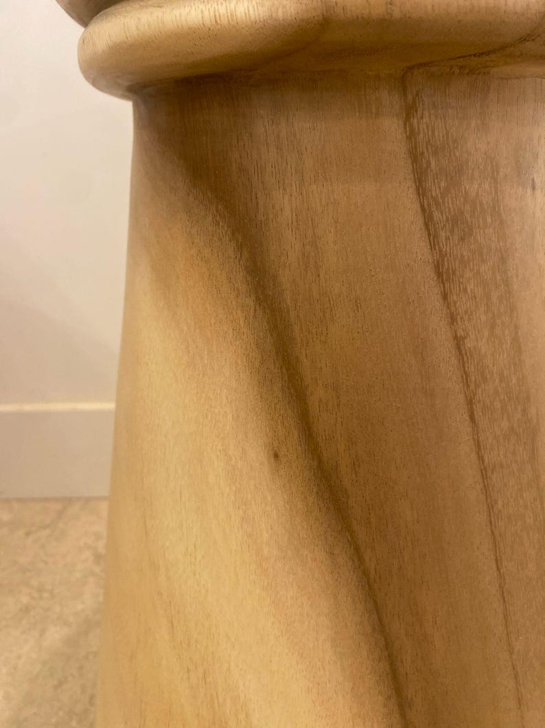 May include: A light brown wooden stool with a smooth, polished finish. The stool features a tapered, cylindrical body with a slightly flared top edge and visible natural wood grain patterns running vertically along the curved surface.