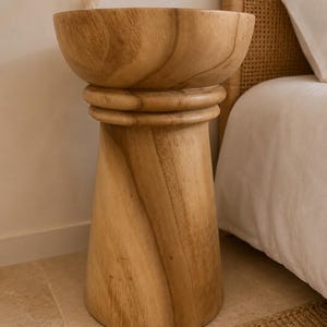 May include: A light brown wooden side table with a smooth, cylindrical base that tapers slightly toward the top. The table features a wide, bowl-shaped top section separated from the base by a thick, rounded horizontal ring detail in the wood.