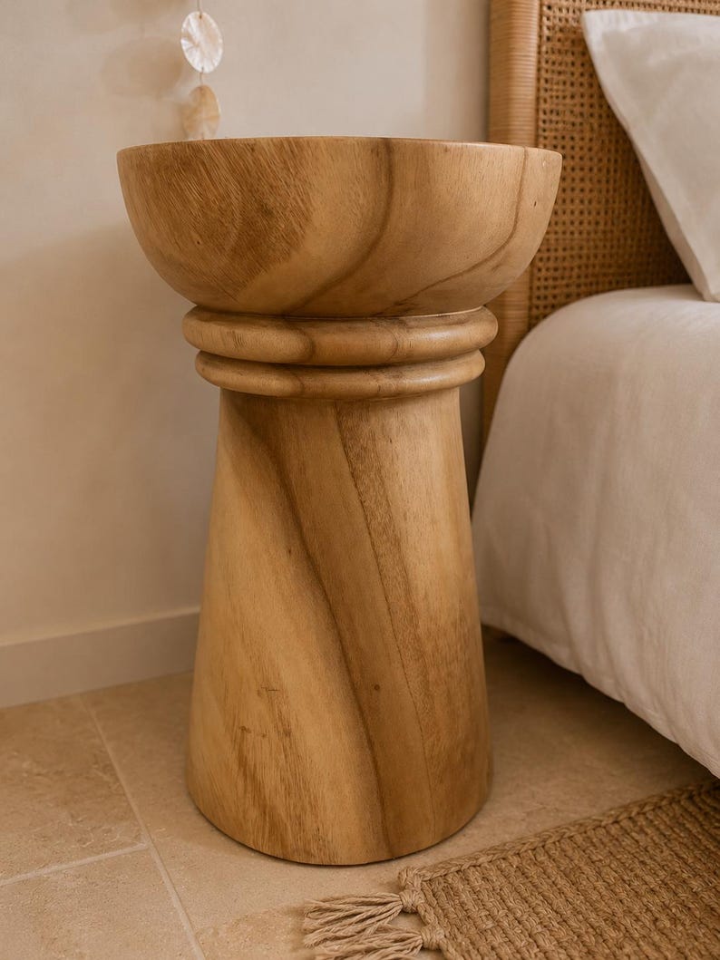 May include: A light brown wooden side table with a smooth, cylindrical base that tapers slightly toward the top. The table features a wide, bowl-shaped top section separated from the base by a thick, rounded horizontal ring detail in the wood.