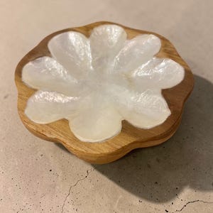 May include: A wooden trinket dish with a flower-shaped design. The dish features a light-colored, pearlescent flower inlay set within a natural wood base. The dish is round with a scalloped edge.