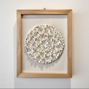 Könnte beinhalten: Ein gerahmtes Kunstwerk mit einer kreisförmigen Anordnung weißer, strukturierter Blumenelemente. Das Kunstwerk befindet sich vor einem neutralen Hintergrund in einem hellen Holzrahmen und erzeugt eine minimalistische Ästhetik.