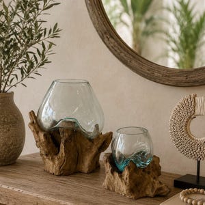 Op de afbeelding: Twee heldere glazen kommen met onregelmatige, golvende randen, elk stevig geplaatst in een natuurlijke houten voet met ruwe randen. De glazen vaten zijn transparant met een lichte blauwe tint, contrasterend met de gestructureerde, donkerbruine houtnerf.