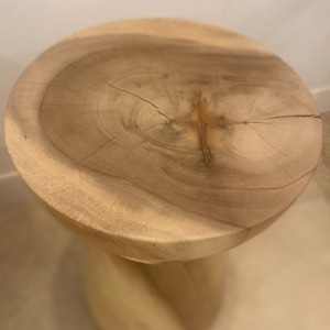May include: A light brown wooden stool featuring a circular top with a visible natural wood grain pattern and a central knot. The base of the stool has a carved, spiraling cylindrical design, showcasing the raw texture and light tan color of the wood.