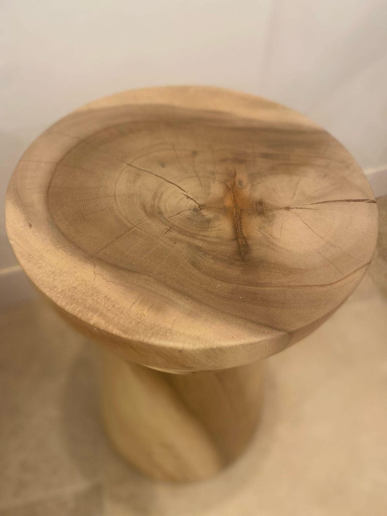 May include: A light brown wooden stool featuring a circular top with a visible natural wood grain pattern and a central knot. The base of the stool has a carved, spiraling cylindrical design, showcasing the raw texture and light tan color of the wood.