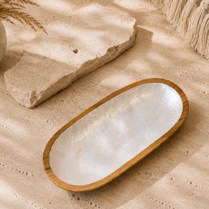 Könnte beinhalten: Ein ovales Tablett mit einem Rand aus Naturholz und einer glatten, schillernden weißen Innenschale. Die flache Schale hat einen sauberen, geschwungenen Rand und eine reflektierende, perlmuttartige Oberfläche, die einen Kontrast zum warmen Holz bildet.