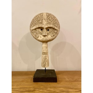 Op de afbeelding: Een gebeeldhouwde houten sculptuur van een gestileerd gezicht met een ronde kop en een lange nek. Het gezicht heeft gedetailleerde kenmerken, waaronder ogen, een neus en een mond. De kop is versierd met geometrische patronen. De sculptuur is gemonteerd op een zwarte rechthoekige basis.