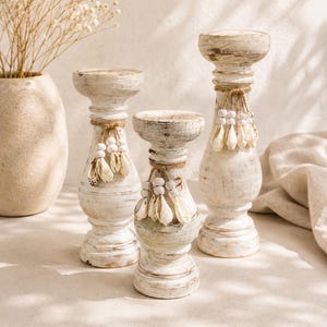 Peut inclure: Trois bougeoirs en bois blanc à l'aspect vieilli de hauteurs variées, chacun doté d'un sommet cylindrique, d'une base en piédestal incurvée et d'une cordelette en jute ornée de petites perles en bois blanc et de coquillages naturels.