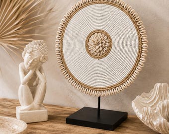 Puede incluir: Una pieza de arte mural circular montada sobre un soporte de metal negro. El disco presenta un centro tejido en blanco, un borde de cuerda color canela y un borde exterior decorado con pequeñas conchas de cauri blancas. Un círculo de cauris decora el centro.