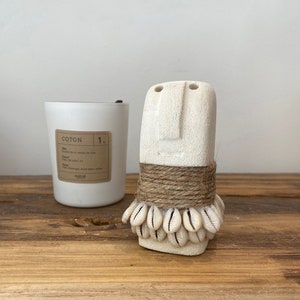 Peut inclure: Sculpture en céramique blanche d'un visage avec une corde et des coquillages autour du cou. La sculpture est posée sur une surface en bois à côté d'une bougie blanche avec une étiquette qui dit "COTON 1."