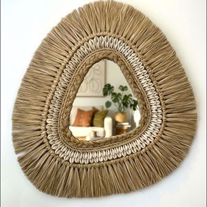 May include: A decorative wall mirror with a unique, organic shape. The frame is made of natural-colored woven fibers, fringed around the edges. The mirror is accented with a braided border and small white seashells, creating a bohemian aesthetic.