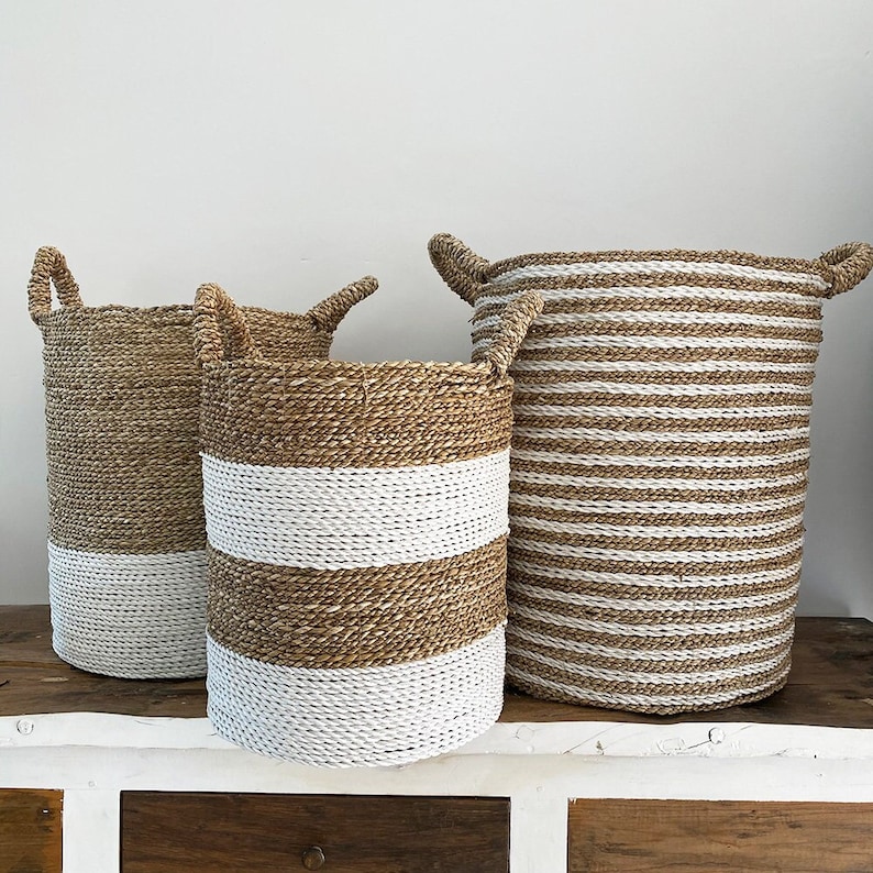 May include: Three cylindrical woven baskets with handles. The left basket is natural tan with a white base. The middle basket features alternating white and natural tan horizontal stripes. The right basket has thin white and natural tan stripes.