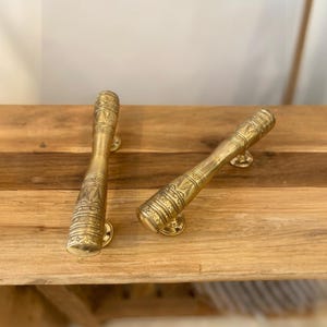 Könnte beinhalten: Zwei Türgriffe aus Messing mit aufwendigen Ätzmustern. Die zylindrischen Griffe haben ausgestellte Enden und sind auf kleinen, runden Sockeln montiert. Sie liegen auf einer Holzoberfläche und unterstreichen ihre dekorative Wirkung.