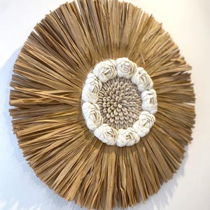 Pode incluir: Decoração de parede redonda com um design central de pequenas conchas claras. O centro é rodeado por um anel de detalhes brancos em forma de concha. A parte externa é feita de fibras vegetais secas, castanho claro, radiantes, criando um efeito de sol.