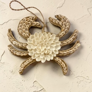 Pode incluir: Enfeite decorativo de caranguejo feito de conchas. O corpo do caranguejo apresenta um design central em forma de flor em branco sujo, rodeado por pernas e pinças bege. Uma argola de barbante é anexada para pendurar. Para decoração costeira.