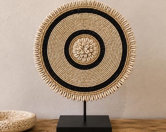 Puede incluir: Una pieza de arte mural circular montada sobre un soporte de metal negro, con anillos concéntricos de fibra natural tejida en tonos beige y negro. El centro y el borde exterior están decorados con conchas de cauri blancas en patrón radial.