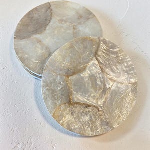 Può includere: Due sottobicchieri rotondi dall'aspetto perlescente e marmorizzato. I sottobicchieri hanno un bordo bianco e una palette di colori naturali e iridescenti con sfumature di crema, beige e oro. I sottobicchieri sono realizzati in un materiale che ricorda la madreperla.