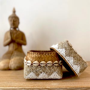 May include: A woven basket with a lid, decorated with small white beads and seashells. The basket has a brown woven interior and a geometric pattern on the exterior. The lid is partially open, revealing the interior.