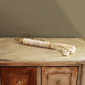 Può includere: Un fiocco bianco e beige fatto di materiali naturali. Il fiocco è fatto di una stringa di perline rotonde bianche con una perlina di legno in cima. Il fiocco è appeso a una superficie di legno.