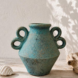 Puede incluir: Un jarrón de cerámica turquesa con un acabado texturizado y desgastado, con dos asas de doble bucle a cada lado. El recipiente presenta un cuerpo ancho y cónico con un cuello estrecho y una abertura circular, con una paleta de colores azul y marrón.